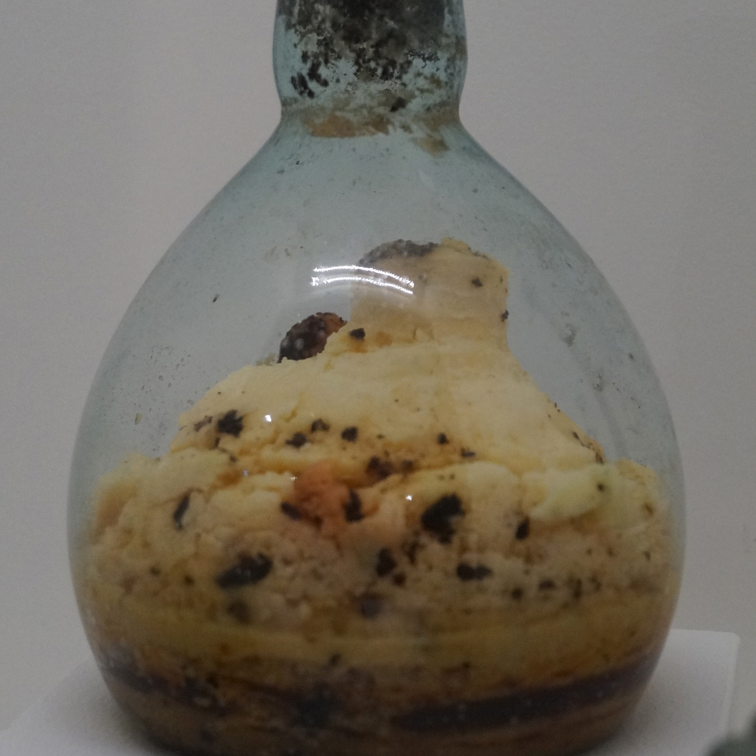Foto einer bauchigen Salbflasche mit schmalem Flaschenhals aus Glas mit verklumptem Inhalt, gefunden als Grabbeigabe. Modern verplombt. Römisch-Germanisches Museum Köln.