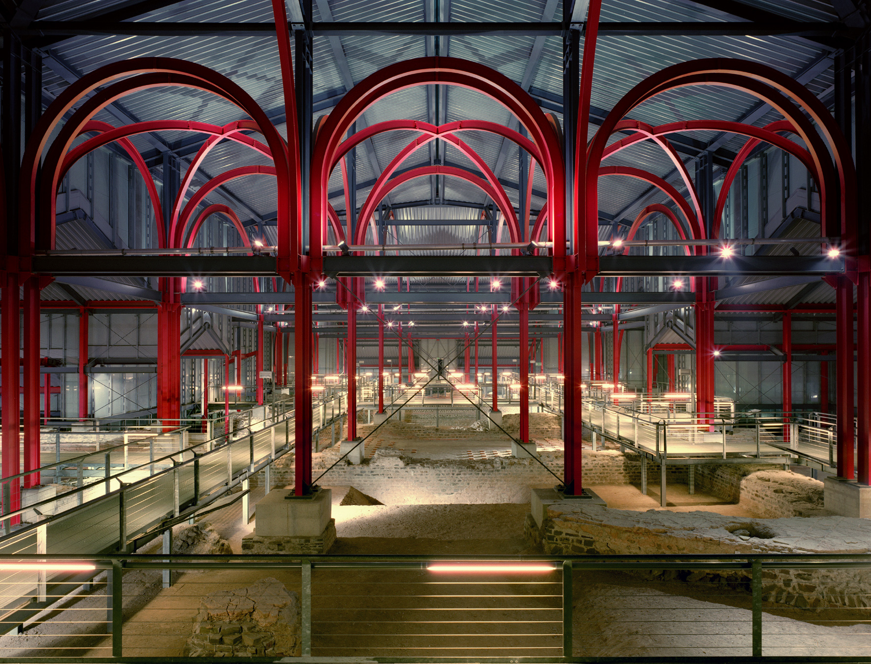 Fotografie des gläsernen Schutzbaus über den großen Thermen im LVR-Archäologischen Park Xanten. Rote Stahlträger tragen die Konstruktion und stehen an den Stellen, wo einst Pfeiler, Wände und Gewölbe waren.