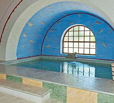 Ein eckiges Wasserbecken in der rekonstruierten Römertherme Carnuntum. Im Hintergrund ein großes, mit Holzstreben  unterteiltes Rundbogenfenster. Die Wände sind aquamarinblau und mit Fischen bemalt.